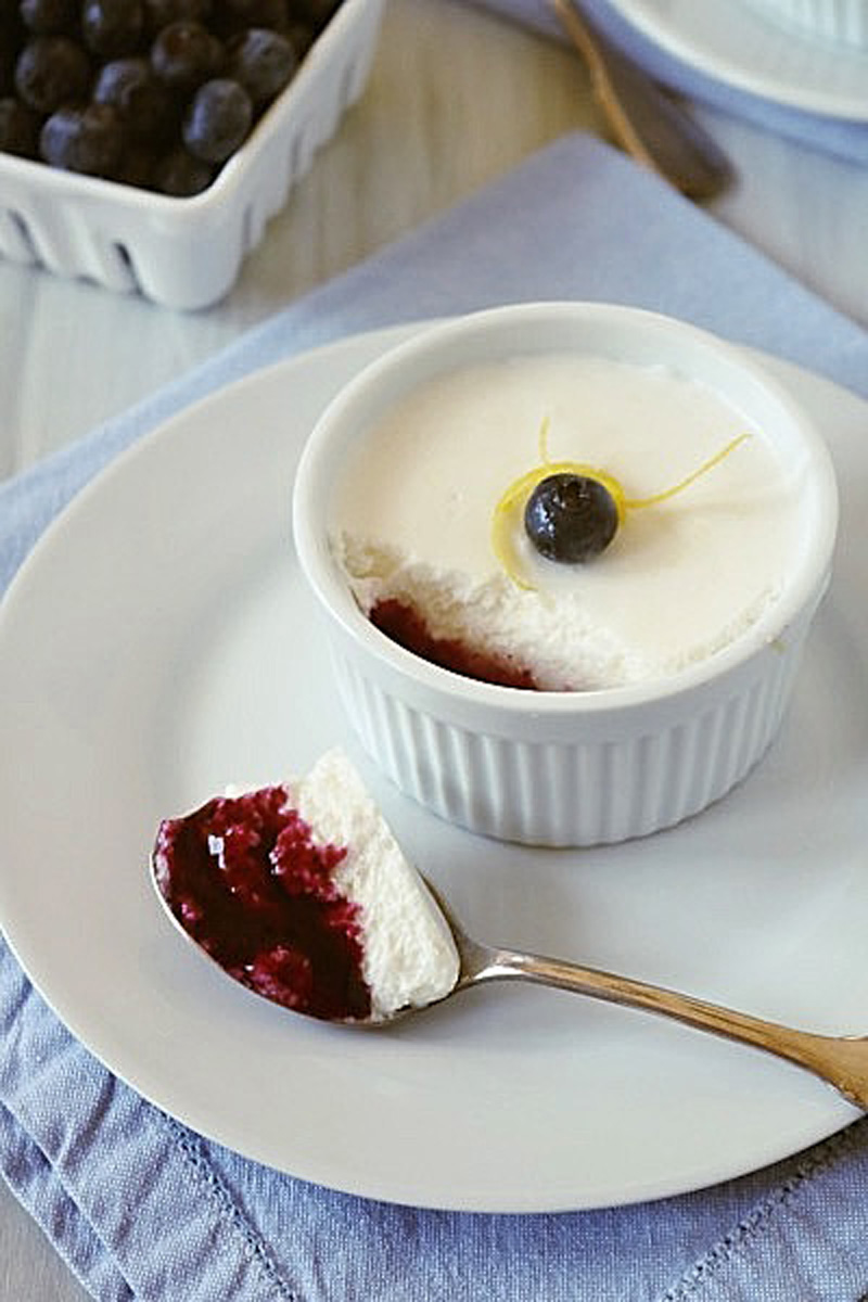 A ramekin of creamy dessert topped with a blueberry and lemon twist sits on a white plate. A spoonful of the dessert, showing a layer of berry filling, rests beside it. Fresh blueberries are in the background.