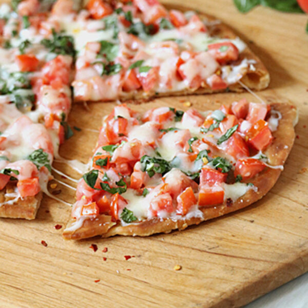 A thin-crust pizza topped with melted cheese, chopped tomatoes, and fresh basil sits on a wooden cutting board. One slice is slightly pulled away from the rest, showing the toppings and crispy crust.