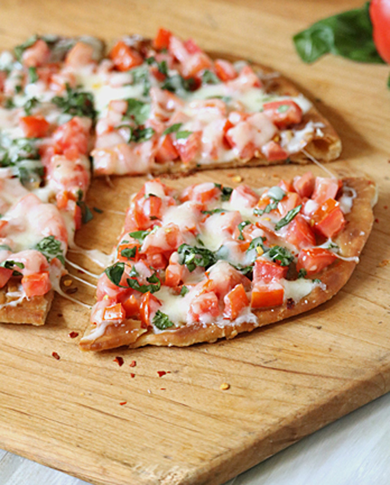 A thin-crust pizza topped with melted cheese, chopped tomatoes, and fresh basil sits on a wooden cutting board. One slice is slightly pulled away from the rest, showing the toppings and crispy crust.