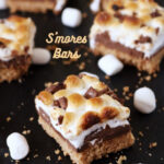 Close-up of S’mores bars with toasted marshmallows, melted chocolate, and a graham cracker crust, arranged on a dark surface with marshmallows scattered around.
