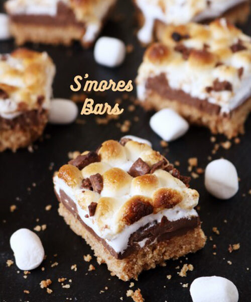 Close-up of S’mores bars with toasted marshmallows, melted chocolate, and a graham cracker crust, arranged on a dark surface with marshmallows scattered around.