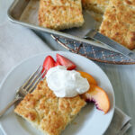 A plate with a square piece of golden-brown biscuit topped with whipped cream, sliced strawberries, and peach slices. A pan with more biscuit squares is in the background. A fork rests on the plate.