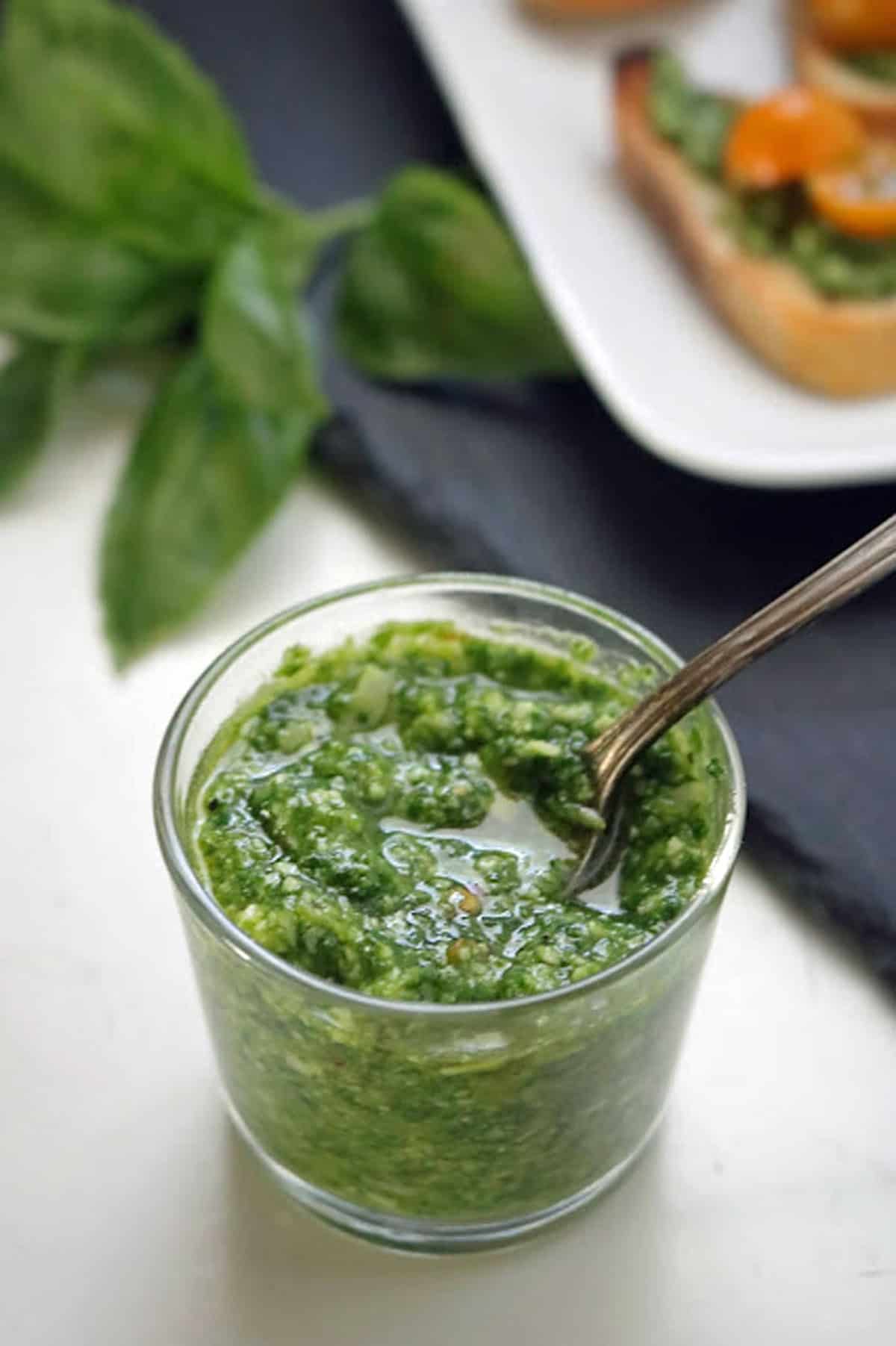 A small glass jar of green pesto with a spoon inside, set on a white surface with fresh basil leaves nearby and slices of bread topped with pesto in the background.