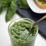 A small glass jar filled with green pesto sauce, with a spoon inside. Fresh basil leaves and a plate of bruschetta are blurred in the background.