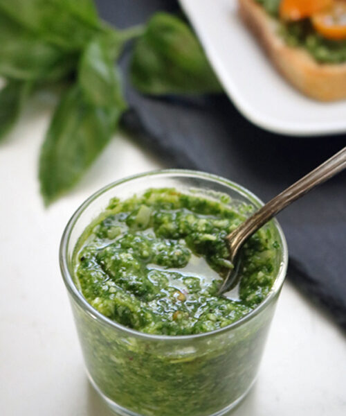 A small glass jar filled with green pesto sauce, with a spoon inside. Fresh basil leaves and a plate of bruschetta are blurred in the background.