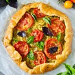 A rustic tomato galette with golden, flaky crust, topped with slices of fresh tomatoes, basil leaves, and herbs, sits on parchment paper. Colorful tomatoes and basil leaves are scattered around it.