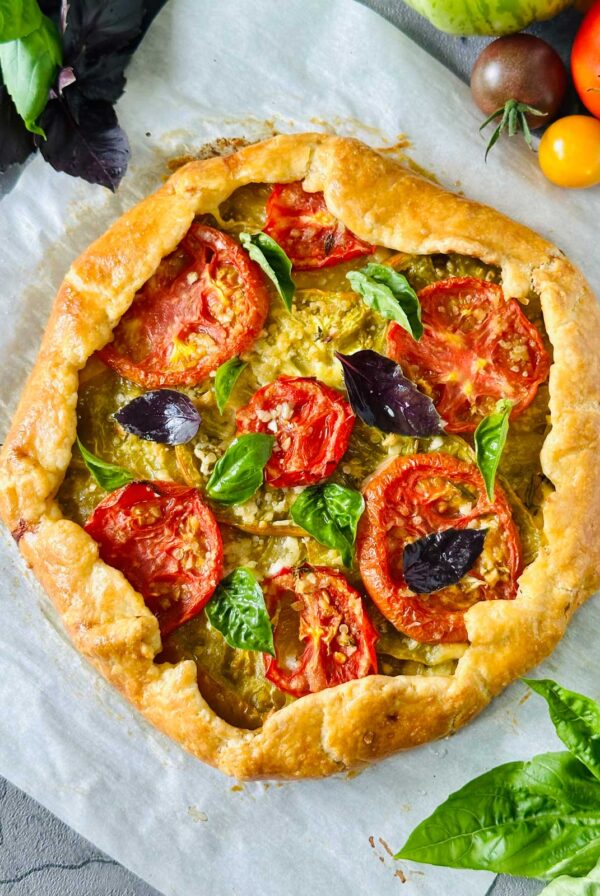 A rustic tomato galette with golden, flaky crust, topped with slices of fresh tomatoes, basil leaves, and herbs, sits on parchment paper. Colorful tomatoes and basil leaves are scattered around it.