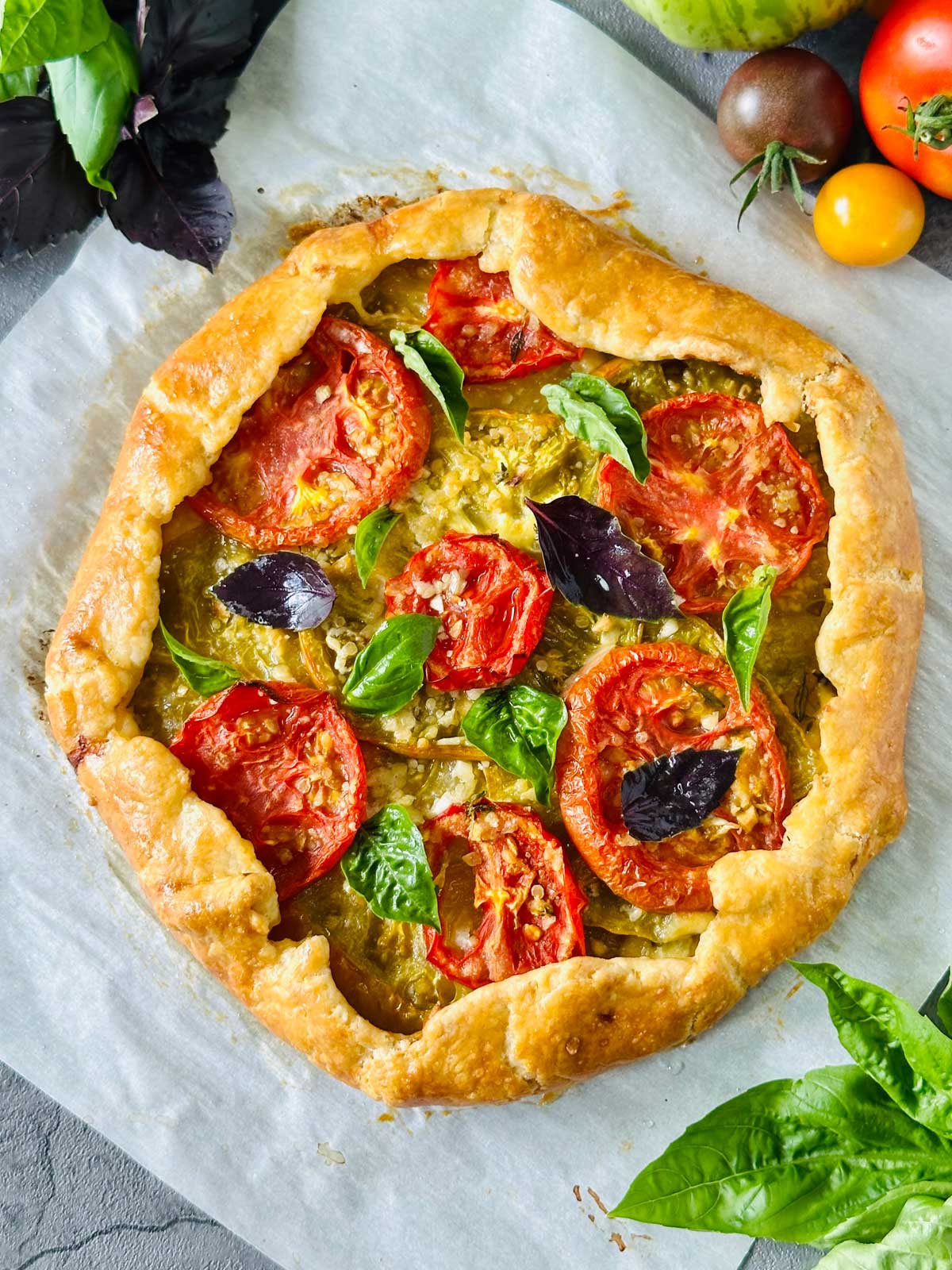 A rustic tomato galette with golden, flaky crust, topped with sliced tomatoes, fresh basil, and purple basil leaves, sits on parchment paper. Colorful tomatoes and basil surround the galette on a gray surface.
