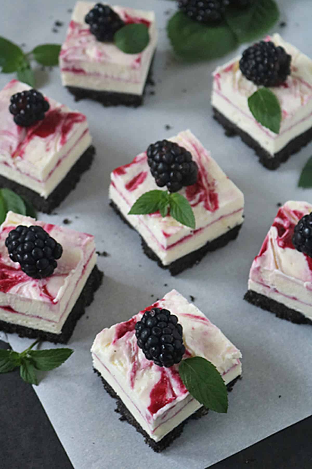Squares of creamy cheesecake with a chocolate crumb crust, topped with a blackberry swirl, fresh blackberries, and mint leaves, arranged on parchment paper.