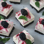 Square cheesecake bars with a chocolate cookie base, swirled berry sauce on top, each garnished with a fresh blackberry and green mint leaves, arranged on parchment paper.