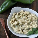 A white bowl filled with a creamy mixture garnished with cilantro sits on a dark wooden surface next to two whole green peppers and a copper spreader knife.