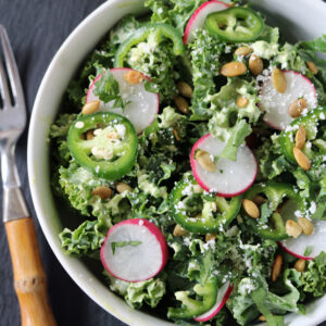A bowl of kale salad topped with sliced radishes, jalapeños, pepitas, crumbled cheese, and fresh herbs is next to a fork with a wooden handle on a dark surface.