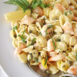 A plate of orecchiette pasta with shrimp, capers, fresh basil, and lemon wedges, served with a spoon on a white plate.
