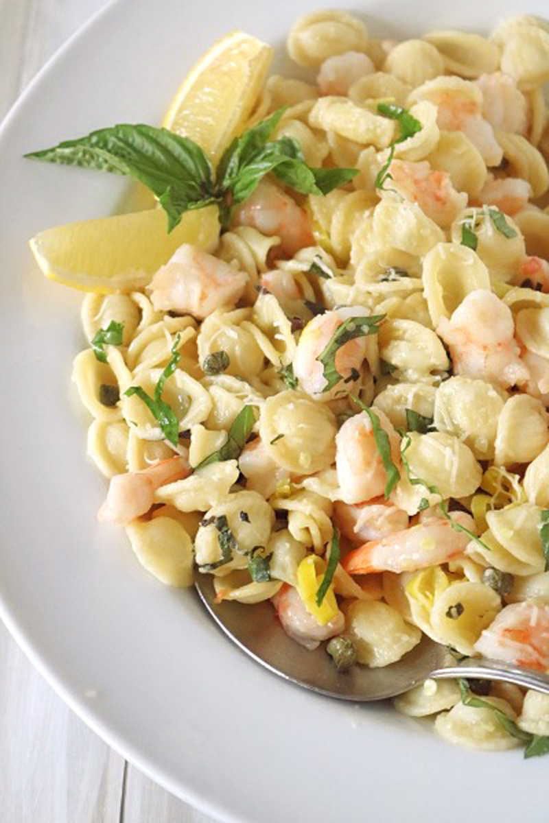 A plate of orecchiette pasta with shrimp, capers, fresh basil, and lemon wedges, served with a spoon on a white plate.