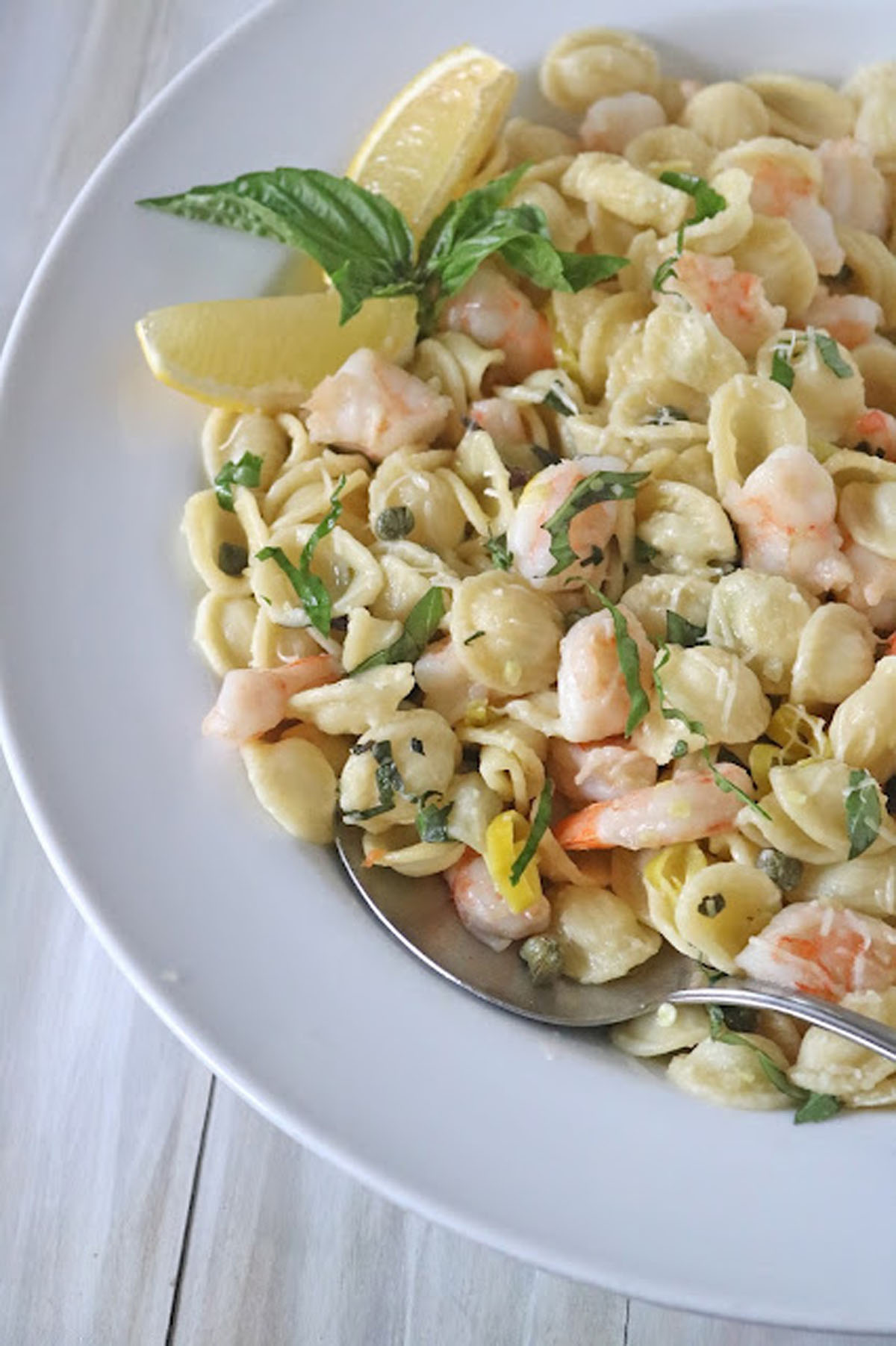 Orecchiette pasta with shrimp, capers, and fresh herbs, garnished with lemon wedges and basil, served on a white plate with a spoon.