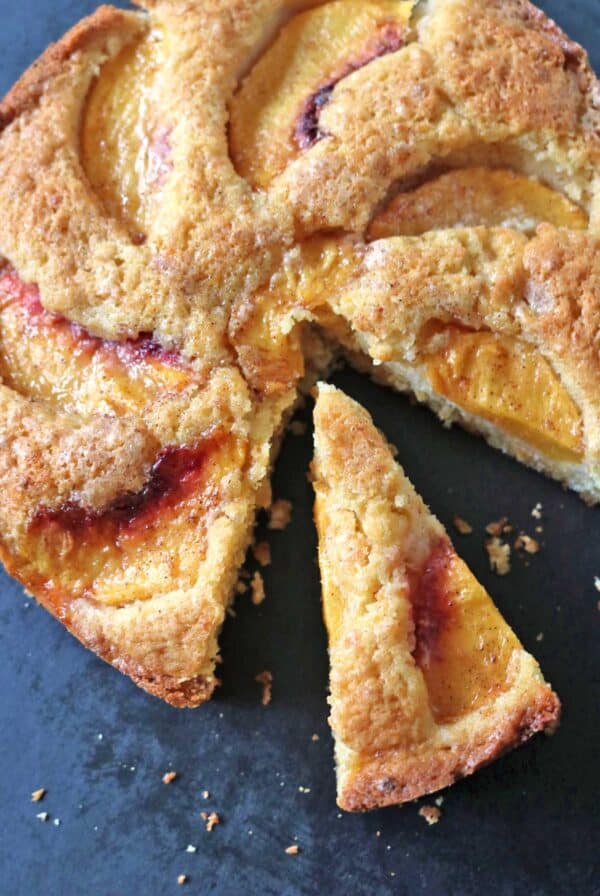 A round peach cake with golden crust, topped with peach slices. One triangular slice has been cut and slightly pulled out from the cake, showing its moist texture. The cake sits on a dark surface.