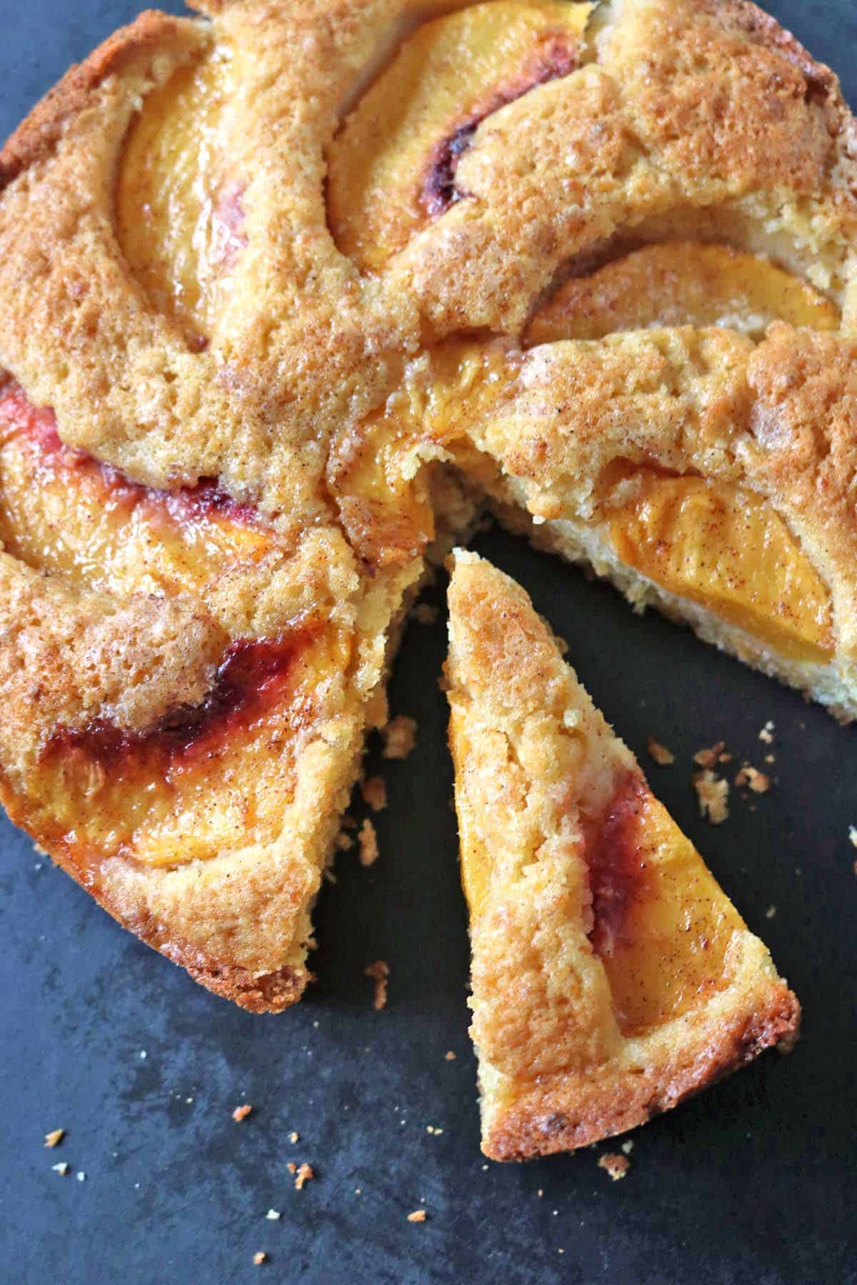A round peach cake with golden crust, topped with peach slices. One triangular slice has been cut and slightly pulled out from the cake, showing its moist texture. The cake sits on a dark surface.