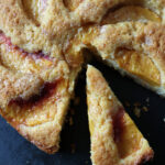 A golden-brown peach cake with a slice cut out, showing juicy peach slices baked into the moist, crumbly cake on a dark surface.