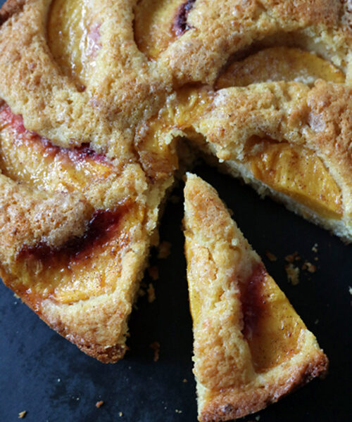 A golden-brown peach cake with a slice cut out, showing juicy peach slices baked into the moist, crumbly cake on a dark surface.