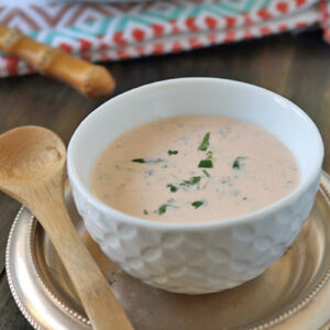 A white bowl filled with creamy pink salad dressing, garnished with chopped herbs, sits on a gold plate with a wooden spoon next to it. A colorful napkin and a plate of leafy greens are in the background.