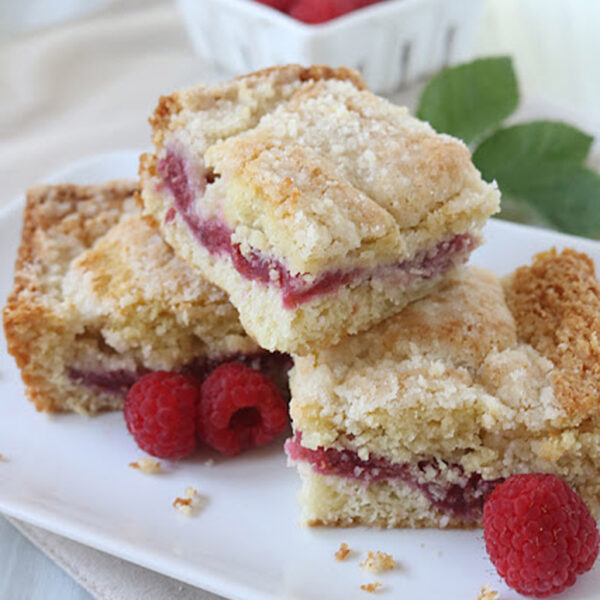 Three raspberry crumb bars are stacked on a white plate, with fresh raspberries scattered around. A white container filled with more raspberries and green leaves is in the background. The bars have a crumbly topping and raspberry filling.