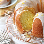 A glazed lemon bundt cake with a slice cut out, displayed on a wire rack with forks nearby. The cake is topped with icing and lemon zest, and a plate with the cut slice is in the background.