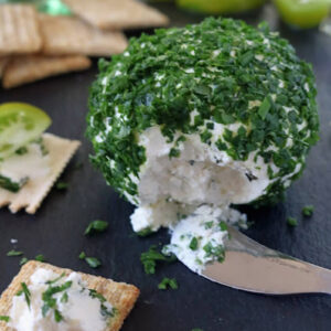 A round cheese ball coated with chopped fresh herbs sits on a dark surface, partially sliced open with a knife. Crackers and green tomato wedges are placed nearby, some topped with the herbed cheese.