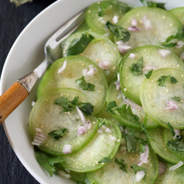 A white bowl contains thinly sliced green tomatillos mixed with chopped onions and fresh cilantro, with a fork resting on the edge. Two whole tomatillos are visible in the background.