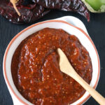 A white bowl filled with red salsa sits on a dark surface, with a wooden spoon inside. Dried red chilies and fresh green tomatillos are placed nearby.