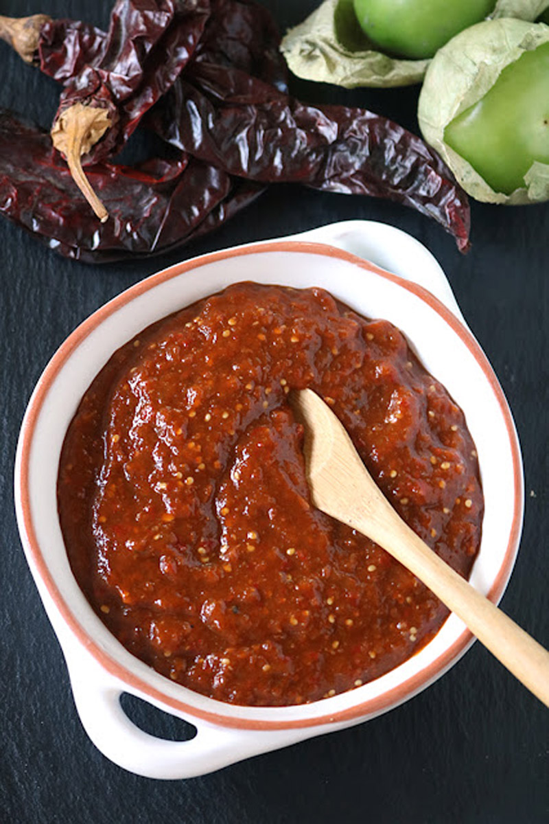 A white bowl filled with red salsa sits on a dark surface, with a wooden spoon inside. Dried red chilies and fresh green tomatillos are placed nearby.