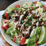 A colorful taco salad in a white bowl topped with lettuce, tomatoes, avocado, black beans, shredded cheese, seasoned ground meat, chopped green onions, and a creamy dressing, with a lime wedge on the side.