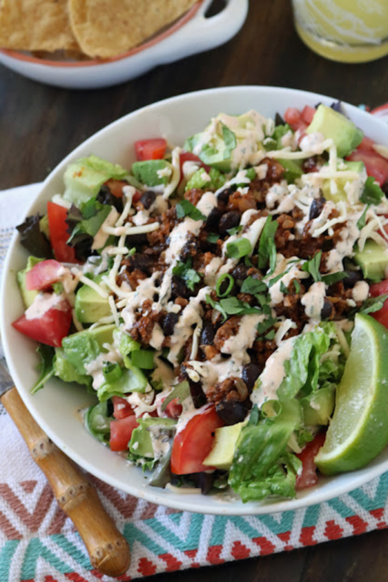 A colorful taco salad in a white bowl topped with lettuce, tomatoes, avocado, black beans, shredded cheese, seasoned ground meat, chopped green onions, and a creamy dressing, with a lime wedge on the side.