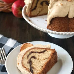 A slice of bundt cake with a cinnamon swirl and creamy frosting sits on a white plate, next to a fork. The rest of the cake, topped with dried apple slices, is displayed on a stand with apples in the background.