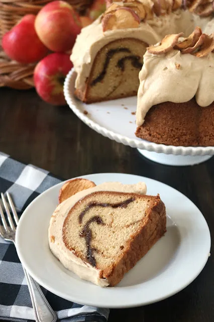 A slice of bundt cake with a cinnamon swirl and creamy frosting sits on a white plate, next to a fork. The rest of the cake, topped with dried apple slices, is displayed on a stand with apples in the background.