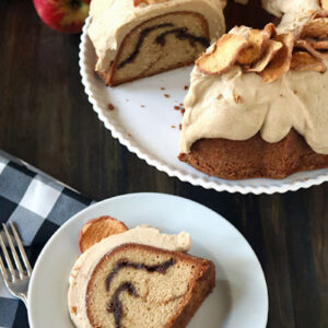 A sliced bundt cake with swirls and cream frosting topped with dried apple slices sits on a white plate; a single slice is served on a smaller plate nearby, next to a fork and a black-and-white checkered napkin.