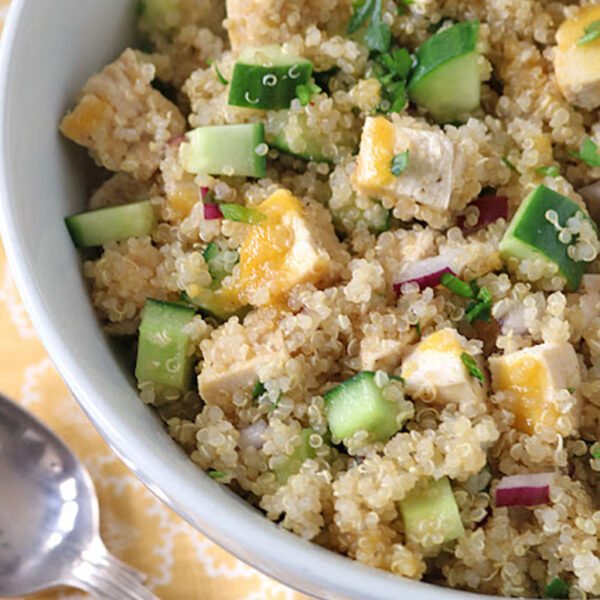 A white bowl filled with quinoa salad containing diced chicken, cucumber, red onion, and fresh herbs, sitting on a yellow-patterned tablecloth next to a silver spoon.