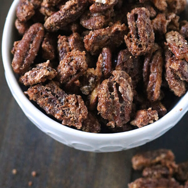 A white bowl filled with candied pecans sits on a dark surface, with a few sugared pecans scattered around the bowl.