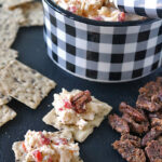 A black-and-white checkered bowl filled with pimento cheese spread, a serving knife, crackers with cheese, and candied pecans arranged on a dark surface.