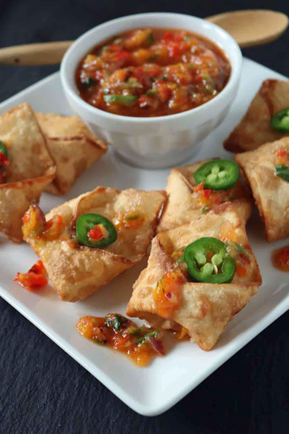 A white plate with crispy wontons topped with sliced jalapeños and sweet chili sauce, served with a bowl of chunky salsa or dip, on a dark surface.