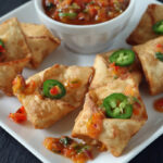 A white plate with crispy wontons topped with sliced jalapeños and colorful salsa, next to a small bowl of salsa. A wooden spoon rests behind the bowl.