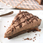 A slice of chocolate mousse pie with a chocolate drizzle sits on a white plate; the pie has a crumbly chocolate crust and caramel oozing out from the side. The remaining pie is on a wire rack in the background.