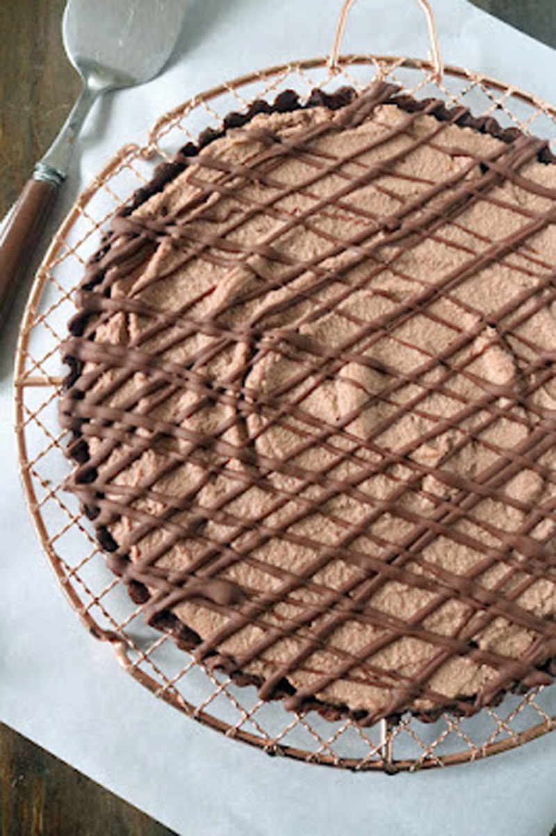 A chocolate tart with a creamy filling and a chocolate drizzle, displayed on a wire cooling rack with a serving spoon beside it.