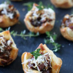 Mini puff pastry cups filled with sautéed mushrooms and topped with grated cheese and fresh herbs, arranged on a dark slate surface.