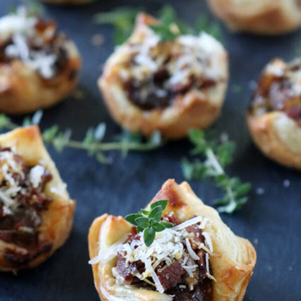 Mini puff pastry cups filled with sautรฉed mushrooms and topped with grated cheese and fresh herbs, arranged on a dark slate surface.