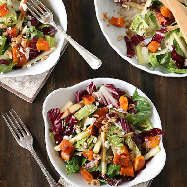Three plates of colorful salad with leafy greens, roasted butternut squash, apple slices, and walnuts, placed on a dark wood table with silver forks and linen napkins; wooden salad servers rest on one plate.