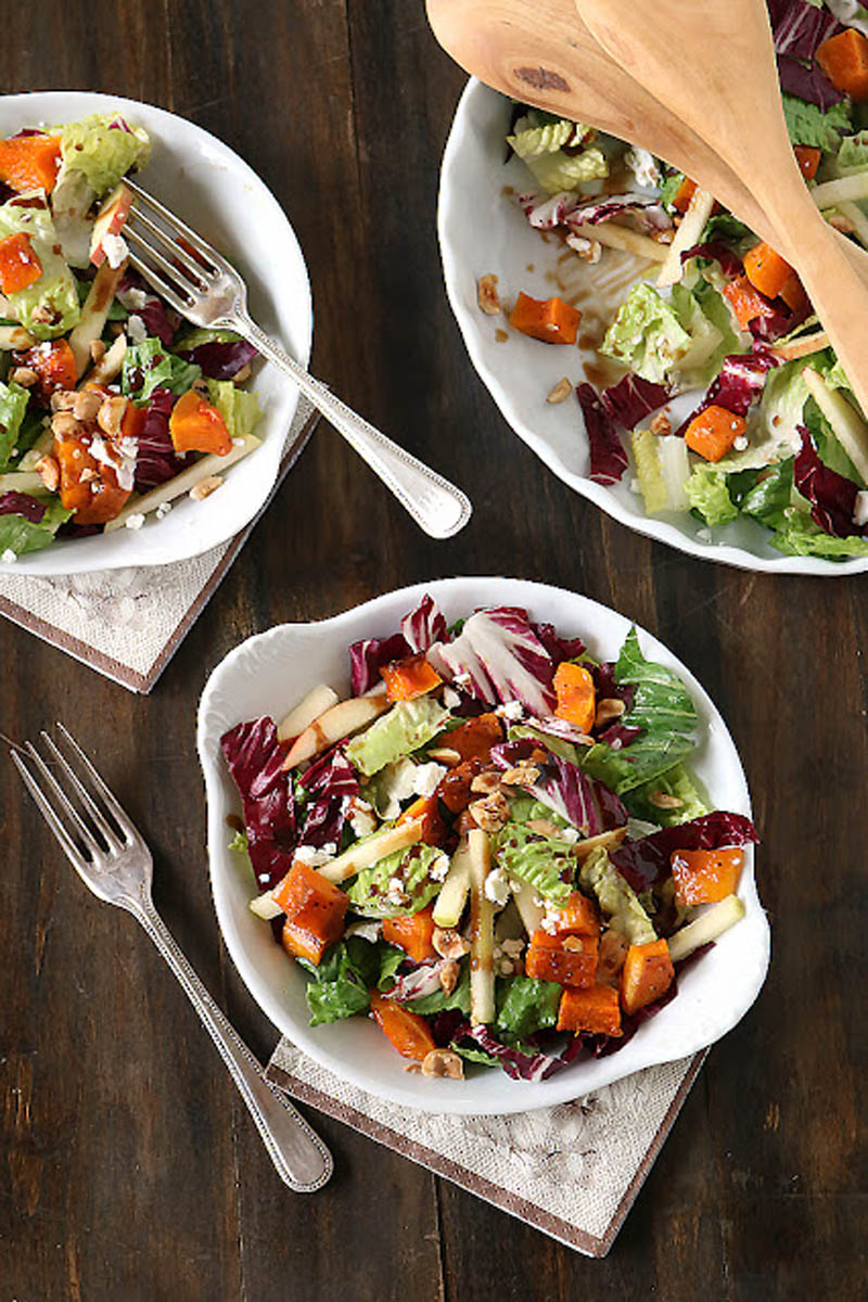 Three plates of colorful salad with leafy greens, roasted butternut squash, apple slices, and walnuts, placed on a dark wood table with silver forks and linen napkins; wooden salad servers rest on one plate.