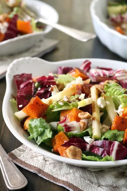 A close-up of a fresh salad in a white bowl, featuring leafy greens, red radicchio, roasted butternut squash, sliced apples, crumbled cheese, and chopped nuts, with another bowl blurred in the background.