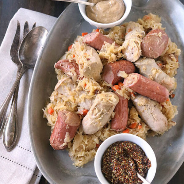 An oval platter with sausages and sauerkraut, accompanied by two small bowls of mustard, set on a dark table with a stack of bread slices, napkins, and silverware nearby.