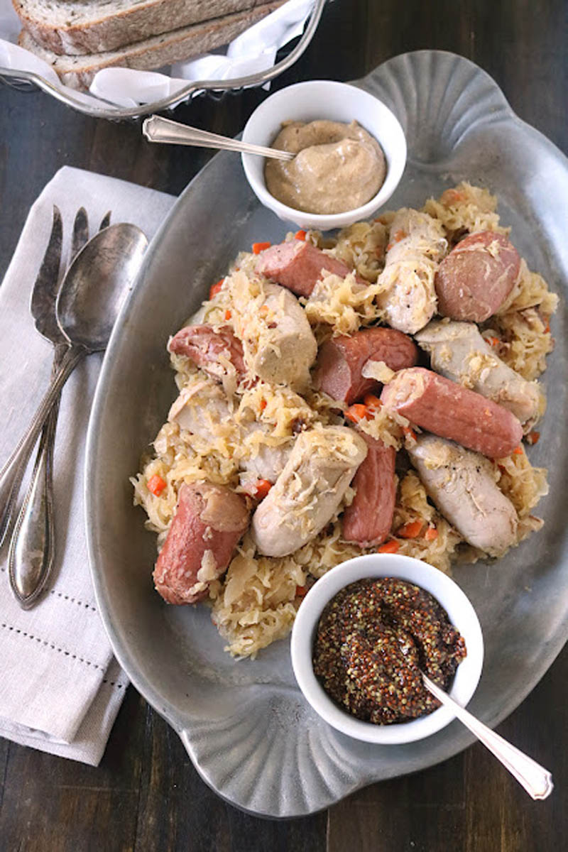 An oval platter with sausages and sauerkraut, accompanied by two small bowls of mustard, set on a dark table with a stack of bread slices, napkins, and silverware nearby.