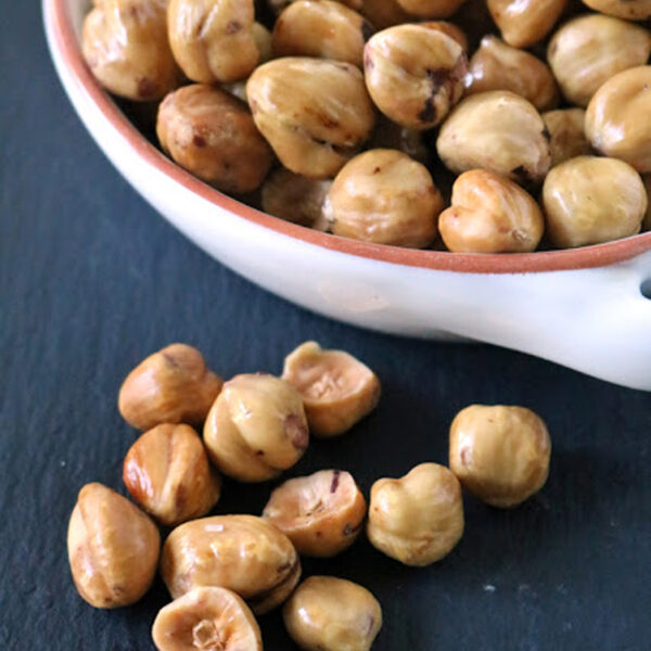A white bowl filled with roasted hazelnuts sits on a dark surface, with several hazelnuts scattered in front of the bowl.
